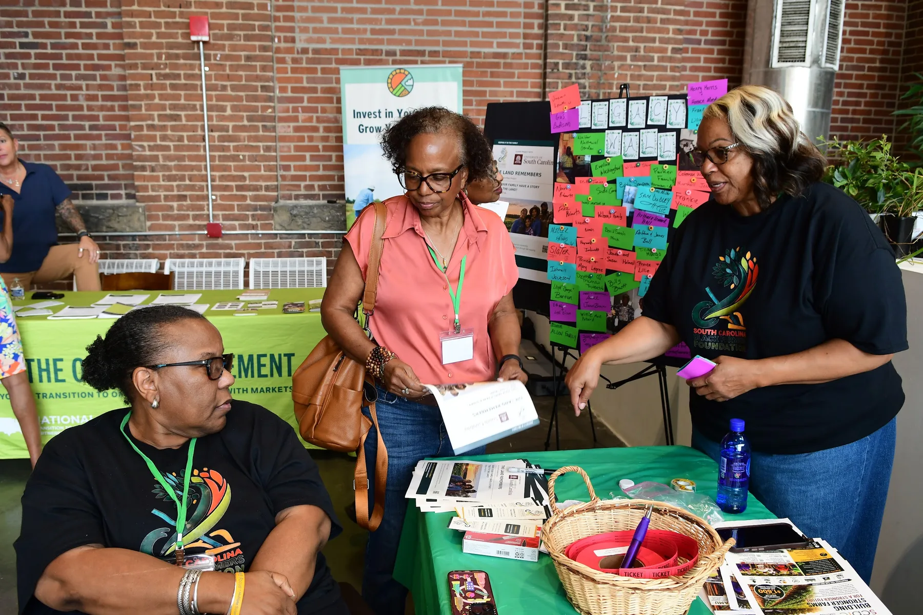 Three women look over printed materials at an information table beside a wall of handwritten notes.