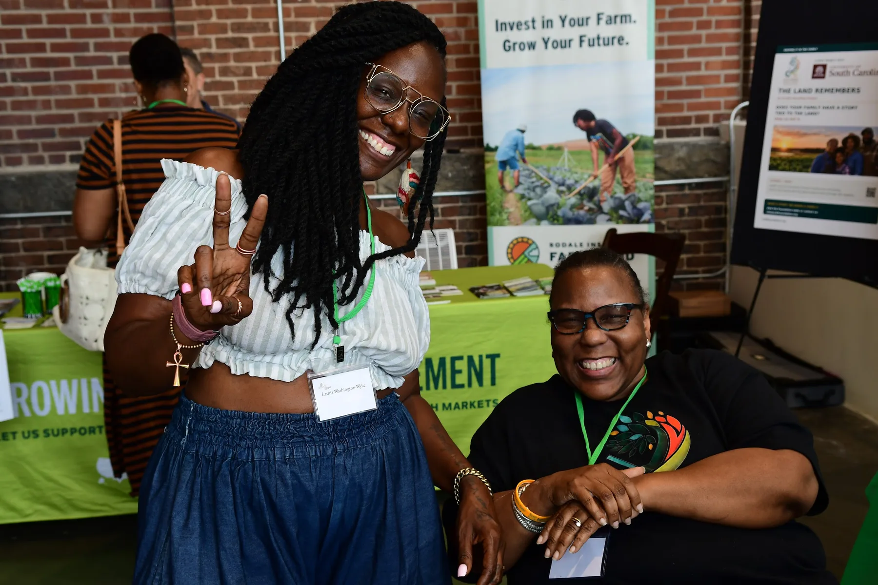Two women smile beside an information table, one flashing a peace sign.